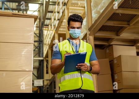 Junge indische Fabrik Lagerarbeiter trägt eine schützende Gesichtsmaske während der Arbeit in der Logistikbranche innen. 30s Mann prüft Artikel Lagerauftrag während Coronavirus Covid 19 Pandemie Ausbruch Stockfoto