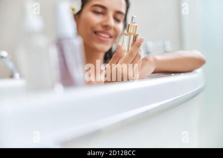 Lächelnde junge Frau hält Flasche Gesicht Serum während der Einnahme Bad Stockfoto