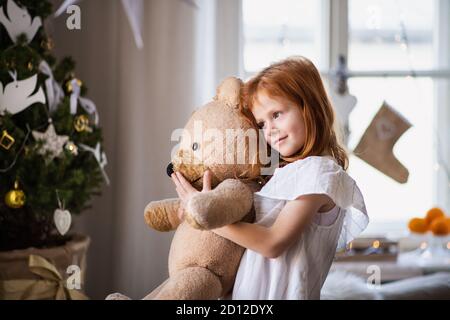 Kleines Mädchen, das zu Weihnachten zuhause steht und Teddybär umarmt. Stockfoto