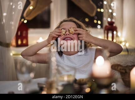 Kleines Mädchen, das zu Weihnachten drinnen sitzt und die Augen mit Lebkuchenkeks bedeckt. Stockfoto