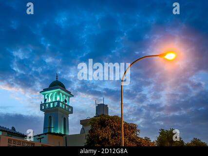 Moschee-Turm in Grün beleuchtet gegen einen blauen Abendhimmel alongwith a hell erleuchteten gelben Straßenlaterne. Aus Maskat, Oman. Stockfoto