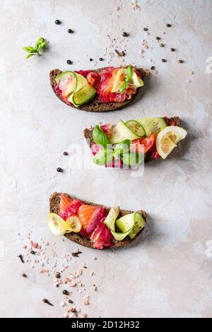 In Scheiben geschnittene rote Beete marinierter Lachs Sandwiches mit Roggen Brot, Gurke, Basilikum und Zitrone über grau Textur Hintergrund. Ansicht von oben, Raum Stockfoto