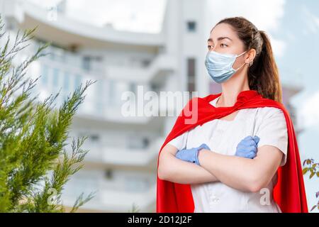 Eine junge Ärztin, die eine OP-Maske und Gummihandschuhe trägt, posiert mit gekreuzten Armen und einem Umhang des Superhelden auf ihrem Rücken. Das Krankenhaus ist verschwommen Stockfoto