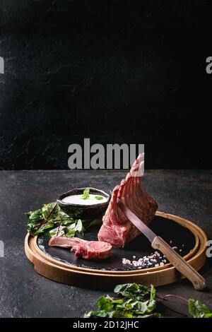 Gegrillter Lammrücken in Scheiben geschnitten mit Joghurt mint Sauce mit grünem Salat junge Rote Bete Blätter, Messer und rosa Salz auf runden hölzernen Schiefer Board serviert über Stockfoto