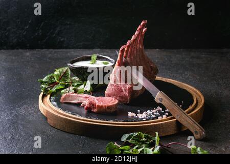 Gegrillter Lammrücken in Scheiben geschnitten mit Joghurt mint Sauce mit grünem Salat junge Rote Bete Blätter, Messer und rosa Salz auf runden hölzernen Schiefer Board serviert über Stockfoto