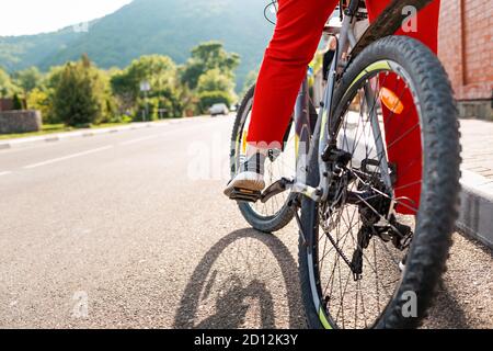 Radfahren. Eine Frau in roter Sportbekleidung sitzt auf einem Fahrrad mit dem Fuß auf dem Pedal. Die Beine sind aus der Nähe. Speicherplatz kopieren. Rückansicht der Straße. Konzept o Stockfoto