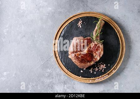 Gegrilltes schwarzes angus-Rind Tomahawk-Steak auf Knochen serviert mit Salz, Pfeffer und Rosmarin auf runder Holzschiefer Schneidebrett auf grauem Textur Hintergrund Stockfoto