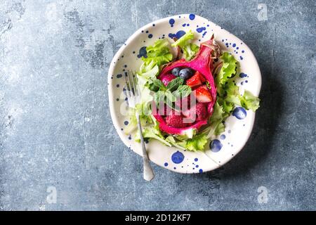 Veganer Obstsalat mit Beeren, grünem Salat und Minze serviert in rosa Drachenfrucht auf getupftem Keramikteller über blauem Texturhintergrund. Draufsicht, Kopieren Stockfoto