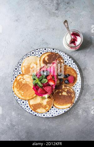 Hausgemachte Pfannkuchen serviert auf dekorieren Teller mit Beeren, Minze und Obstsalat in rosa Drachenfrucht, Glas Joghurt über graue Textur backgrou Stockfoto