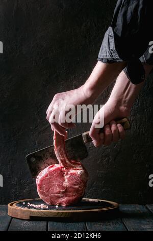 Des Menschen Hände schneiden Roh ungekocht Black Angus Rind tomahawk Steak auf den Knochen von Vintage Metzger Cleaver auf runden Holz- Schiefer Schneidbrett in dunklem Holz Stockfoto