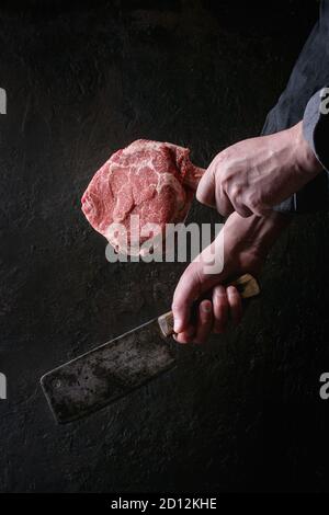 Des Menschen Hände halten Roh ungekocht Black Angus Rind tomahawk Steaks auf Knochen und vintage Metzger Cleaver auf dunklem Hintergrund. Im rustikalen Stil Stockfoto