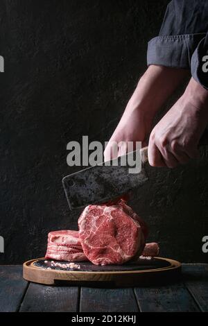 Des Menschen Hände schneiden Roh ungekocht Black Angus Rind tomahawk Steaks auf den Knochen vintage Metzger Cleaver auf runden Holz- Schiefer Schneidbrett über dunkel wird Stockfoto