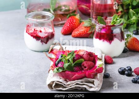Vegan Fruchtsalat mit Beeren und Minze serviert in Pink dragon Frucht auf Tuch mit Gläser Joghurt, eine Flasche Limonade, Zutaten oben auf grau Tabelle. Stockfoto