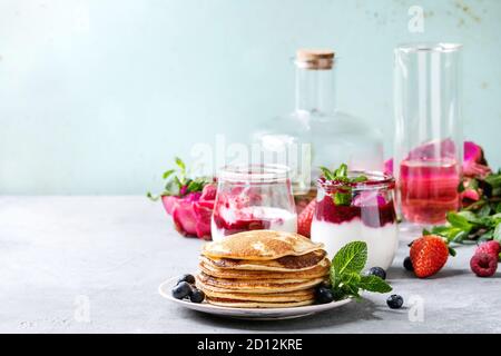 Stapel von hausgemachte Pfannkuchen auf Platte mit Beeren, Minze, Glas Gläser Joghurt, eine Flasche Limonade, Obstsalat in Pink dragon Obst über Graues te Stockfoto