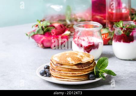 Stapel von hausgemachte Pfannkuchen auf Platte mit Beeren, Minze, Glas Gläser Joghurt, eine Flasche Limonade, Obstsalat in Pink dragon Obst über Graues te Stockfoto