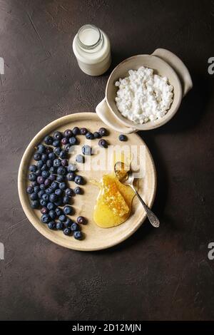 Keramik Schale hausgemachter Hüttenkäse mit Blaubeeren, Flasche Milch und Waben über Dunkelbraun Textur Hintergrund serviert. Ansicht von oben, Raum Stockfoto