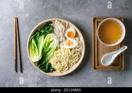 Zutaten zum Kochen asiatische Gericht udon Nudeln mit gekochtem Ei, Pilze, boc Choy, Fleischbrühe serviert in Keramik Schüssel mit Löffel und Stäbchen über Grau Stockfoto