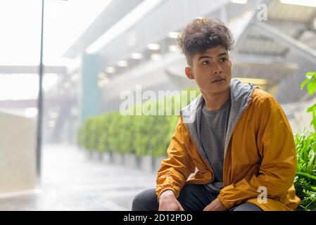 Junge asiatische Mann mit lockigen Haaren denken und sitzen in Die Stadt im Freien mit Regen Stockfoto