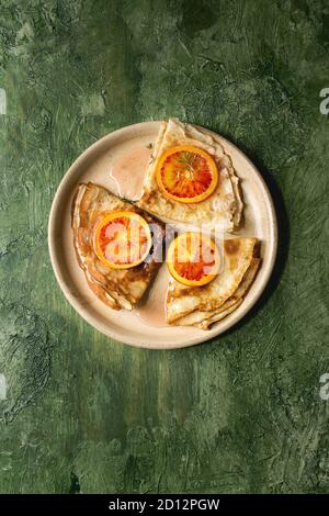 Hausgemachte crepes Pfannkuchen in Keramik Platte mit blutigen Orangen und Rosmarin Sirup mit in Scheiben geschnittenen sizilianischen Rotwein Orangen über grüne Textur Hintergrund serviert. Stockfoto