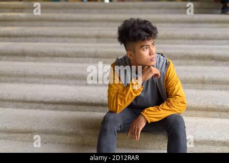 Junge asiatische Mann mit lockigen Haaren denken und sitzen auf Treppe in der Stadt Stockfoto