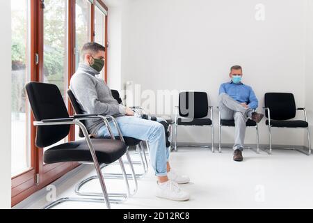 Junger und reifer Mann mit Gesichtsmasken sitzt in einem Wartezimmer eines Krankenhauses oder Büros - konzentrieren Sie sich auf Der junge Mann Stockfoto