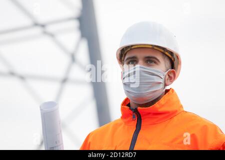 Junger Ingenieur oder Architekt mit Gesichtsmaske und Plan in Seine Hand trägt schützende Arbeitskleidung und Hut - Selektiver Fokus Stockfoto