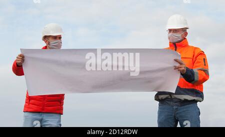 Junge und reife Ingenieure oder Arbeiter diskutieren einen Bauplan Mit Gesichtsmaske und sozialer Distanzierung - Fokus auf die Gesichter Stockfoto