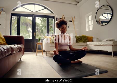 Junge gesunde schöne Frau in sportlichen Top und Leggings üben Yoga zu Hause in Lotushaltung auf Yogamatte sitzend Meditierend lächelnd entspannt mit Stockfoto