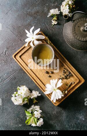 Traditionelle Keramik heiße Tasse grünen Tee auf Holzbrett mit schwarzem Eisen Teekanne, spring flowers White Magnolia und Cherry blühende Zweige über Dunkel Stockfoto