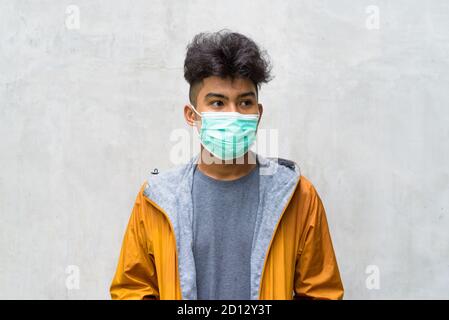 Junge asiatische Mann mit lockigen Haaren trägt Maske und Denken Gegen Betonwände