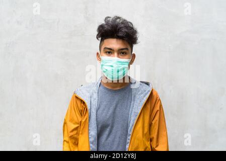 Junger asiatischer Mann mit lockigen Haaren und Maske gegen Beton Wand Stockfoto