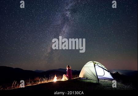 Wanderweibchen entspannt in der Sommernacht Camping in den Bergen neben Lagerfeuer, glühend Touristenzelt. Junge Frau Backpacker genießen Blick auf den Nachthimmel voller Sterne und Milchstraße. Reiseabenteuer-Konzept Stockfoto