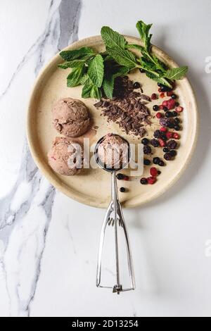 Hausgemachte Schokolade Himbeere Eis Kugeln serviert mit gefrorenen Beeren, Minze, gehackt dunkle Schokolade und Metalllöffel in Keramik Platte über weiße Marb Stockfoto