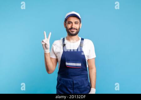 Optimistischer Handwerker in Overalls und Hygienehandschuhe mit Siegeszeichen, Beruf der Dienstleistungsbranche, Kurierdienst, Housekeeping Wartung. W Stockfoto