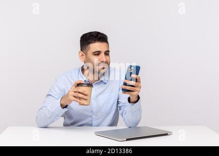 Eleganter, fröhlicher Geschäftsmann, der mit Kaffee in der Hand sitzt und Nachrichten abliest, E-Mails auf dem Handy liest und am Arbeitsplatz eine Pause macht. Stockfoto