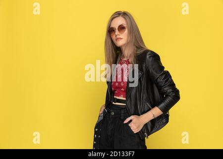 Selbstbewusstes Modelmodell, das beide Hände in den Taschen hält und eine Sonnenbrille trägt, während es auf gelbem Studiohintergrund steht Stockfoto
