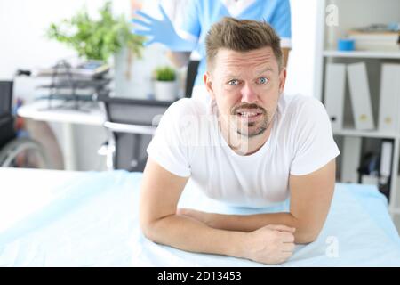 Junger männlicher Patient liegt auf der Couch auf dem Bauch mit Arzt Tragen von Gummihandschuhen im Hintergrund Stockfoto