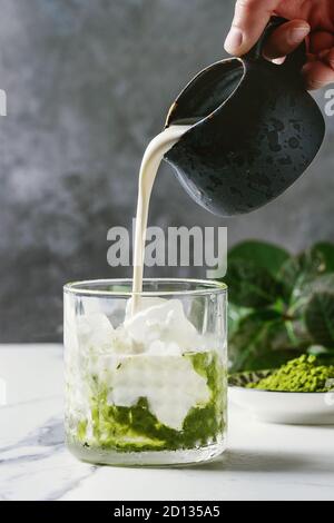 Creme gießen von Krug zu Matcha Grüntee Iced Latte oder Cocktail in Glas mit Eiswürfeln, Matcha Pulver auf weißem Marmortisch, von grünen br eingerichtet Stockfoto