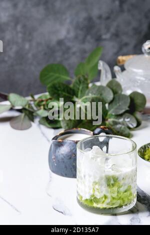Matcha Grüntee Iced Latte oder Cocktail in Glas mit Eiswürfeln, Matcha Pulver und kanne Milch auf weißem Marmor Tabelle, durch grüne Zweige eingerichtet. Gre Stockfoto
