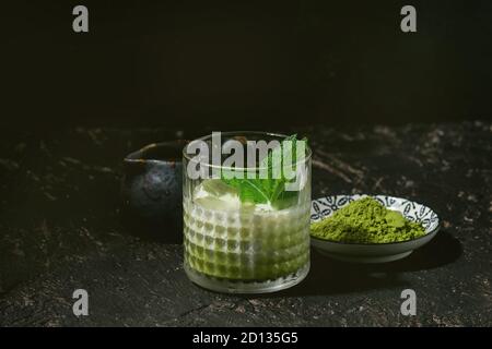 Matcha Grüntee Iced Latte oder Cocktail in Glas mit Eiswürfeln, Minze, Matcha Pulver und kanne Milch über dunkle Textur Hintergrund Stockfoto