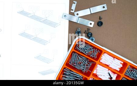 Schrauben und Wandstecker in orangefarbener Kunststoffbox. Werkzeugkasten von oben auf braunem Hintergrund mit Montageanleitung für Spültrockner. Satz Schrauben. Stockfoto