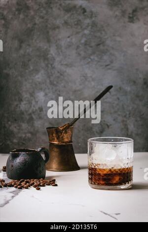 Eiskaffee Cocktail oder Frappe mit Eiswürfeln im Glas mit Vintage jezva, Milchkännchen und Kaffeebohnen auf weißem Marmor tisch mit grauer Beton w Stockfoto