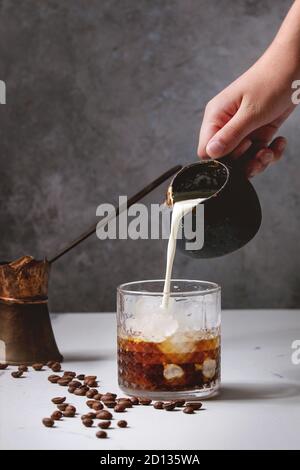 Eiskaffee Cocktail oder Frappe mit Eiswürfeln und Creme, Gießen von Kanne, in Glas mit Vintage jezva und Kaffeebohnen auf weißem Marmor serviert. Stockfoto