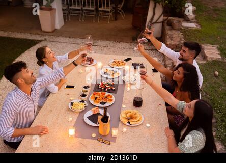 Draufsicht auf Gruppe von Freunden, die mit Rotwein toasten Gläser bei Sonnenuntergang auf einem großen Tisch aus weißem Granit Der Garten mit Köstlichkeiten und brennenden Kerzen beladen Stockfoto