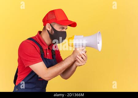Profil Porträt eines Handyman in Unionalls und schützende medizinische Maske mit Lautsprecher, Warnung vor sozialer Distanz und Sicherheit während der Quarantäne. Stockfoto