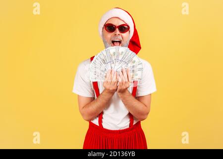 Extrem aufgeregt glücklich grauen bärtigen Mann in weihnachtsmann Hut und stilvolle Sonnenbrille hält Fan von Dollar, Geld an Feiertagen, Boni. Innen s Stockfoto