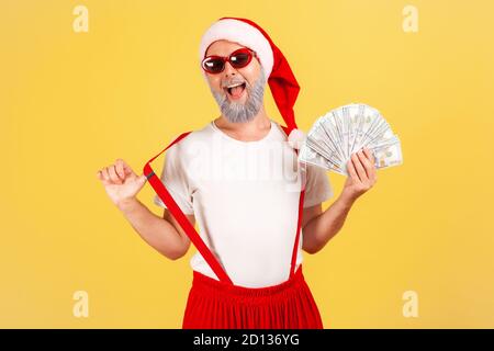 Aufgeregt positive ältere Mann in weihnachtsmann Hut und Sonnenbrille zeigt Fan von Dollar Blick auf die Kamera mit extrem zufrieden Ausdruck, Wetten. Stockfoto