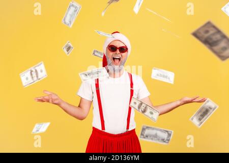 Extrem lächelnd positive Erwachsene Mann in weihnachtsmann Hut und trendige Sonnenbrille genießen unter Dollar Banknoten stehen regen, Lotterie Sieg, Einkommen. Stockfoto