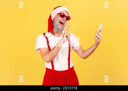 Lächelnder, positiver älterer Mann in weihnachtsmannhut, der Friedensgesten zeigt, der per Videoanruf auf seinem Smartphone spricht, Blogger, der mit Abonnenten kommuniziert Stockfoto