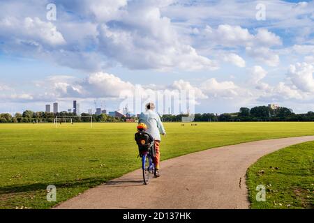 Großbritannien, London, Hackney Marshes. Ein Radfahrer mit einem Kleinkind in einem Sicherheitssitz auf einem Radweg in einem Londoner Park. Sonnig, Sommertag. Stockfoto